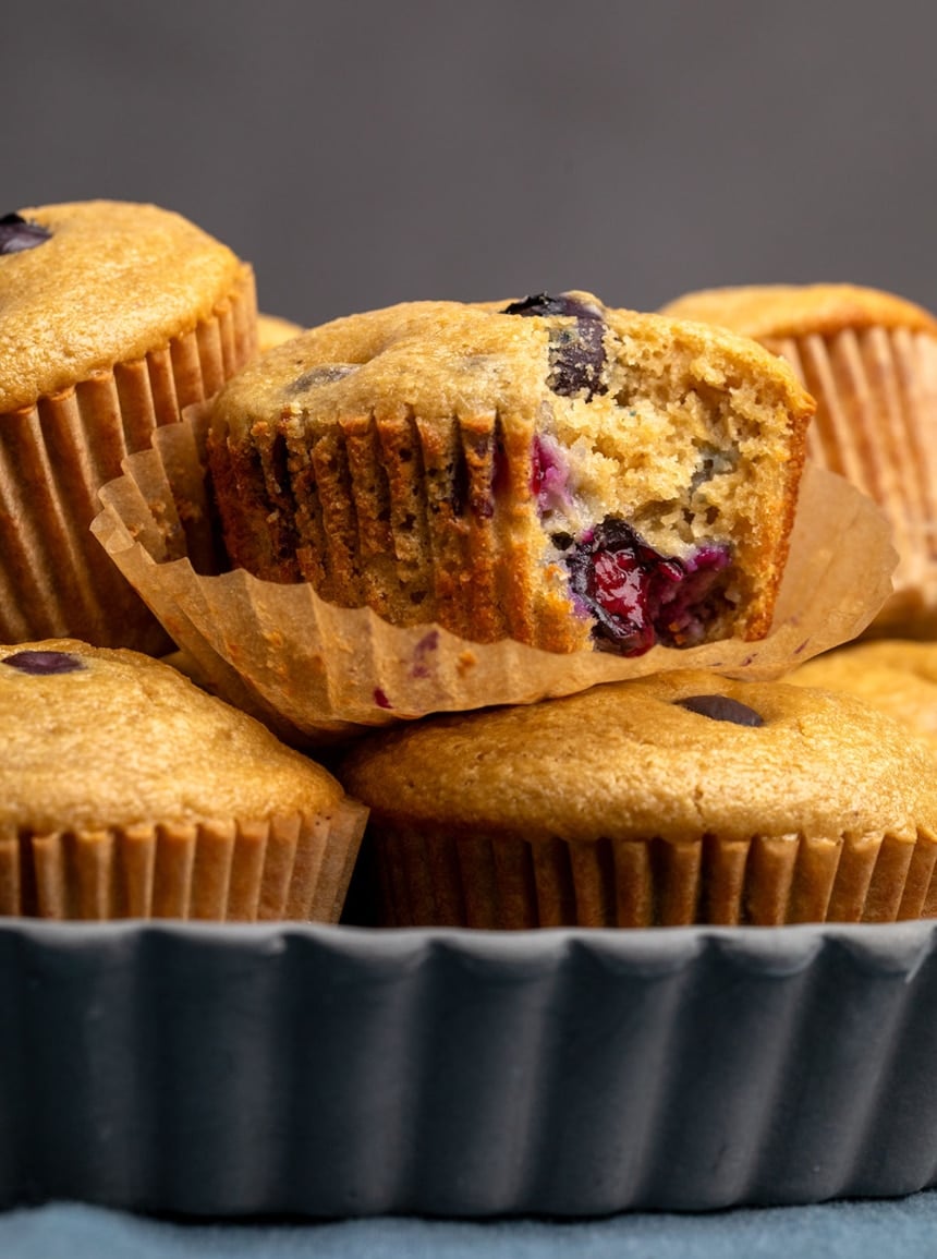 pile of light brown muffins with blueberries baked inside in brown muffin liners in dark gray dish with 1 1/2 inch sides sitting on blue cloth, with one muffin missing a single bite