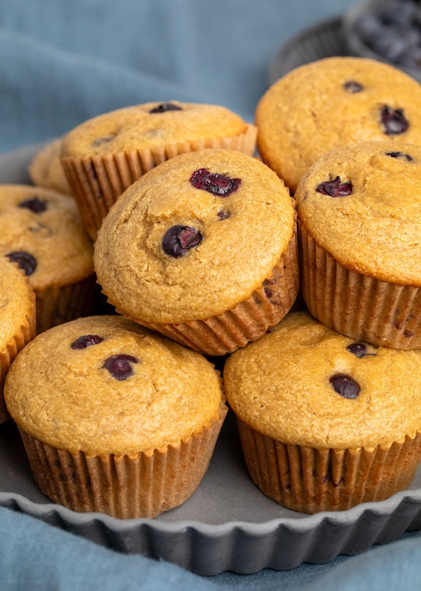 pile of whole light brown muffins with some blueberries peeking out of top of each muffin and on side through brown paper wrapping