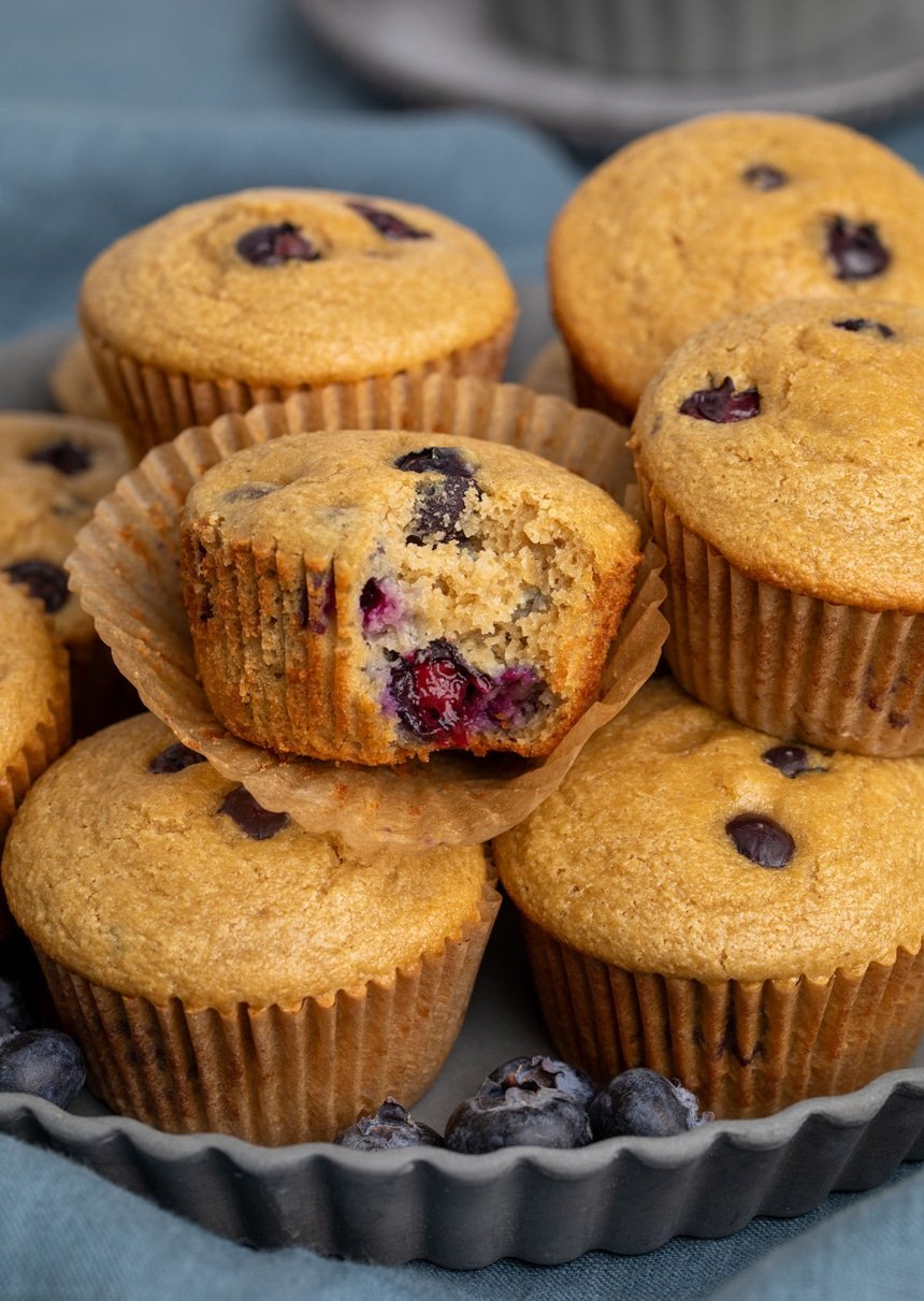 pile of light brown muffins with blueberries in a dark gray round dish with fluted edges with some fresh whole blueberries around the muffins, and one muffin with a bite taken revealing baked bright berries inside