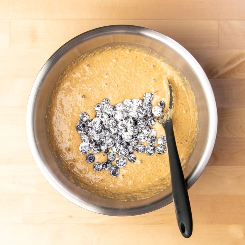 Same mixing bowl with soft light brown raw batter with fresh blueberries on top covered in white cornstarch with a black silicone spatula