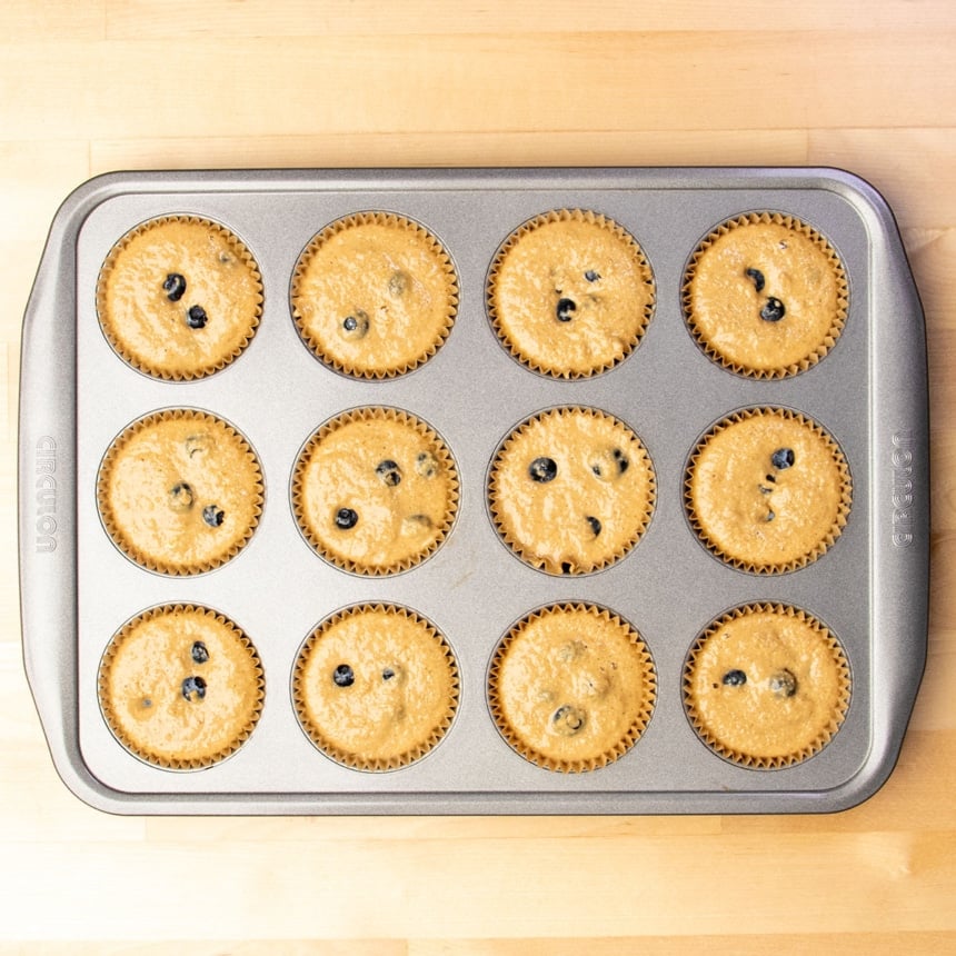 Gray Circulon brand standard 12-cup muffin tin with brown liners in each well filled with light brown raw muffin batter with visible blueberries