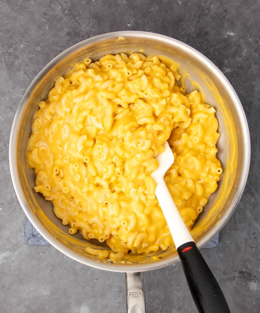 round metal saucier pan with cooked elbow macaroni mixed with light orange-yellow cheese sauce with white silicone spatula with black handle on gray background