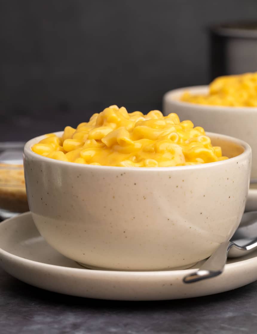 side view of small but tall sand colored bowl on small plate with metal spoon on the plate and elbow macaroni in orange yellow cheese sauce inside