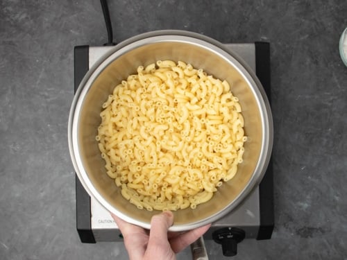 hand holding round medium size metal bowl with cooked light tan elbow macaroni noodles