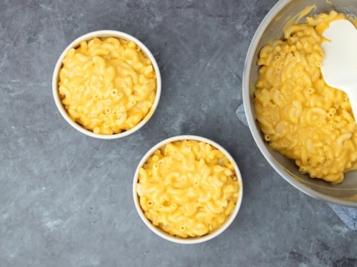 overhead image of 2 small round bowls with elbow macaroni in yellow orange cheese sauce with rest of dish in metal saucepan, all on a gray-black surface