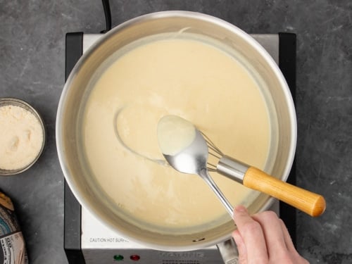 thickened mixture in saucepan with same whisk with fingers holding a large metal spoon turned upside down with sauce clinging to the back of the spoon