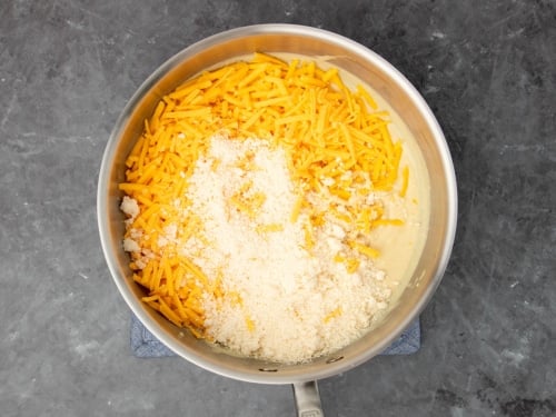 white grated cheese and yellow shredded cheese on top of thickened pale mixture in saucepan