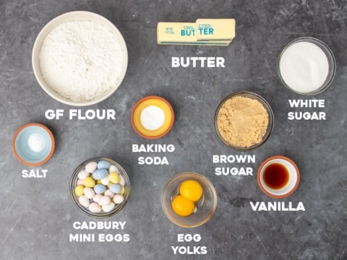 ingredients for gluten free Cadbury egg cookies in small bowls with white block letters with names of ingredients on black marble surface