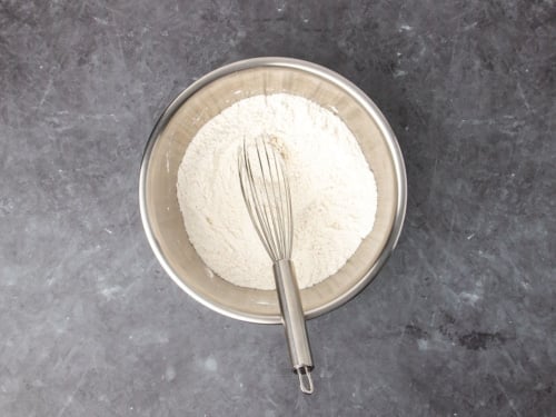 Small round metal mixing bowl with white flour mixture and large metal wire whisk