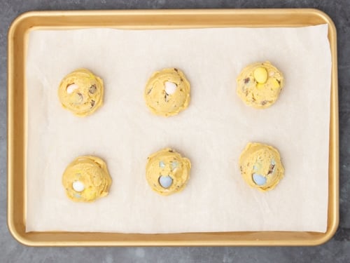 6 mounds of raw light brown cookie dough with whole blue, white, pink and yellow candy pieces pressed into the top on white paper on gold baking tray