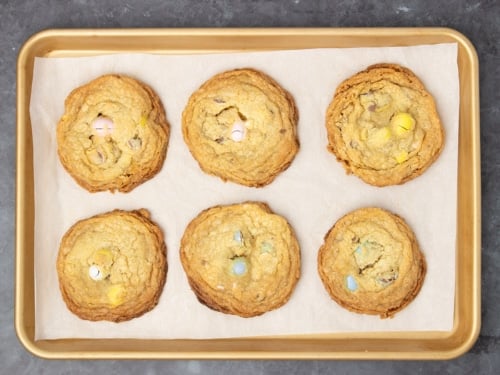 Same baking sheet with 6 mounds of cookie dough baked into 6 golden brown round cookies with darker edges and colored candy pieces peeking through the tops of the cookies