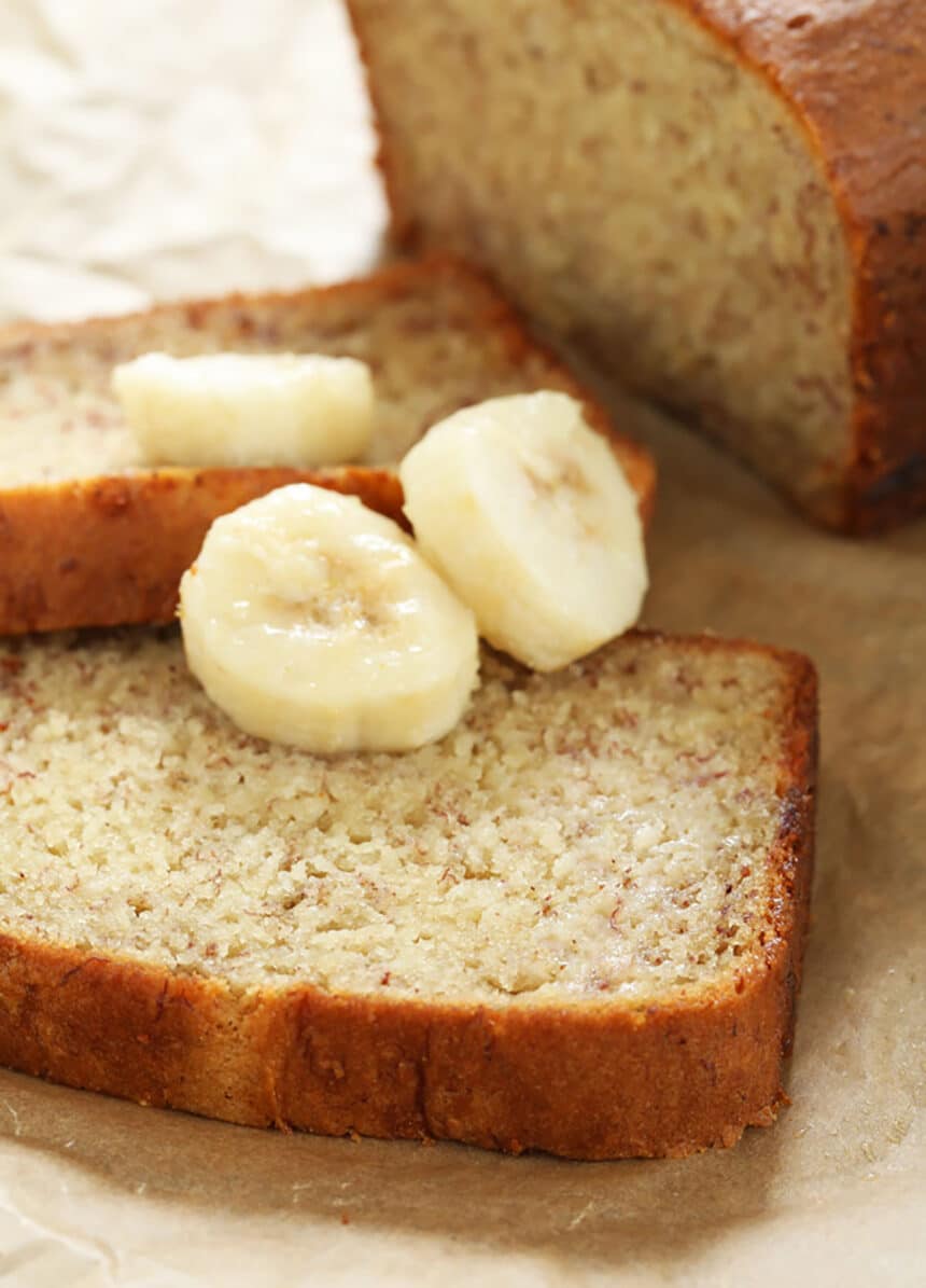 closeup of a slice of light brown gluten free banana bread with 3 slices of fresh bananas on brown paper