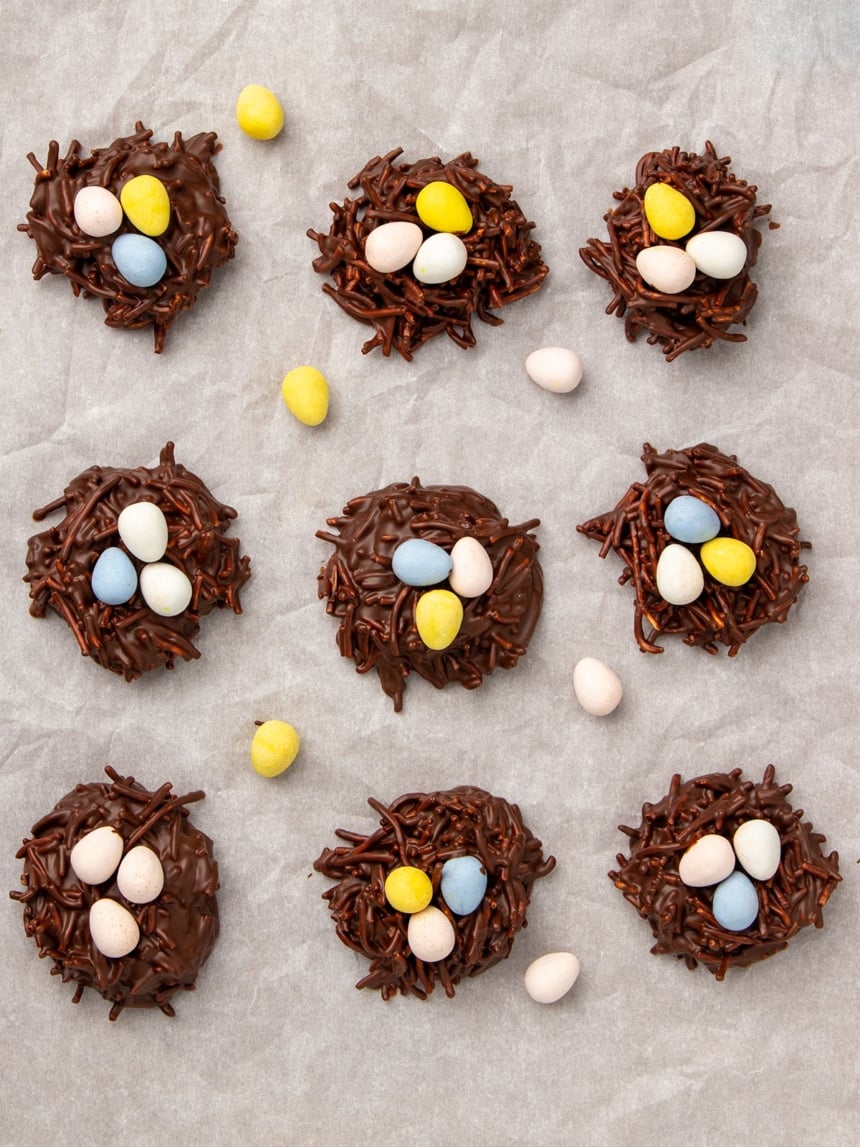 overhead image of 9 chocolate brown no bake cookies that resemble bird's nests with 3 pastel-colored mini egg shaped candies on top of each, all on white paper