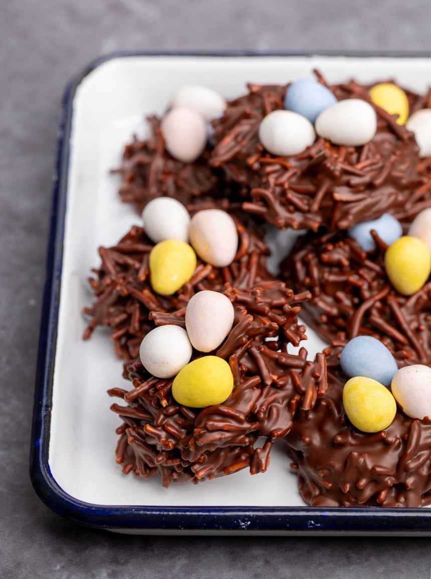 pile of dark brown cookies with chow mein noodles mixed in to melted chocolate and topped with mini pastel egg shaped candies on a small white tray with blue rim