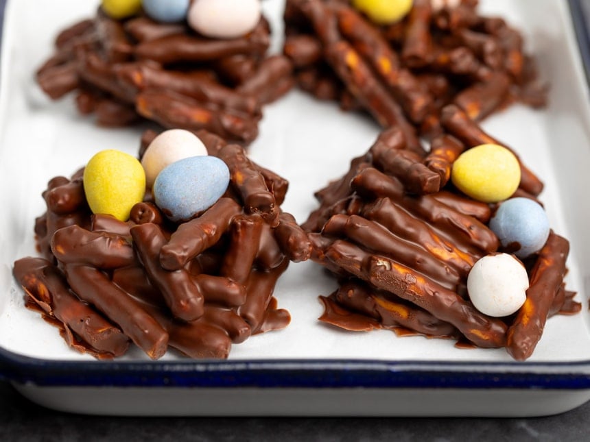 white tray with blue rim with 4 brown bird's nest cookies made with broken pretzel sticks and melted chocolate topped with 3 Cadbury mini egg candies each