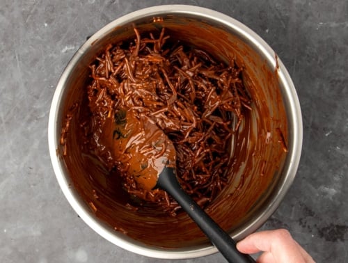 fingers holding black silicone spatula using it to turn over mixture and mix together chow mein noodles with melted chocolate