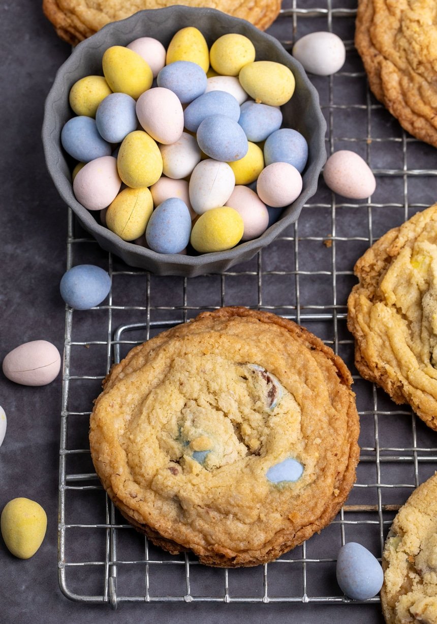 Small gray stone dish with pale yellow, blue pink and white Cadbury mini egg candies on wire rack with golden brown cookies with darker brown edges