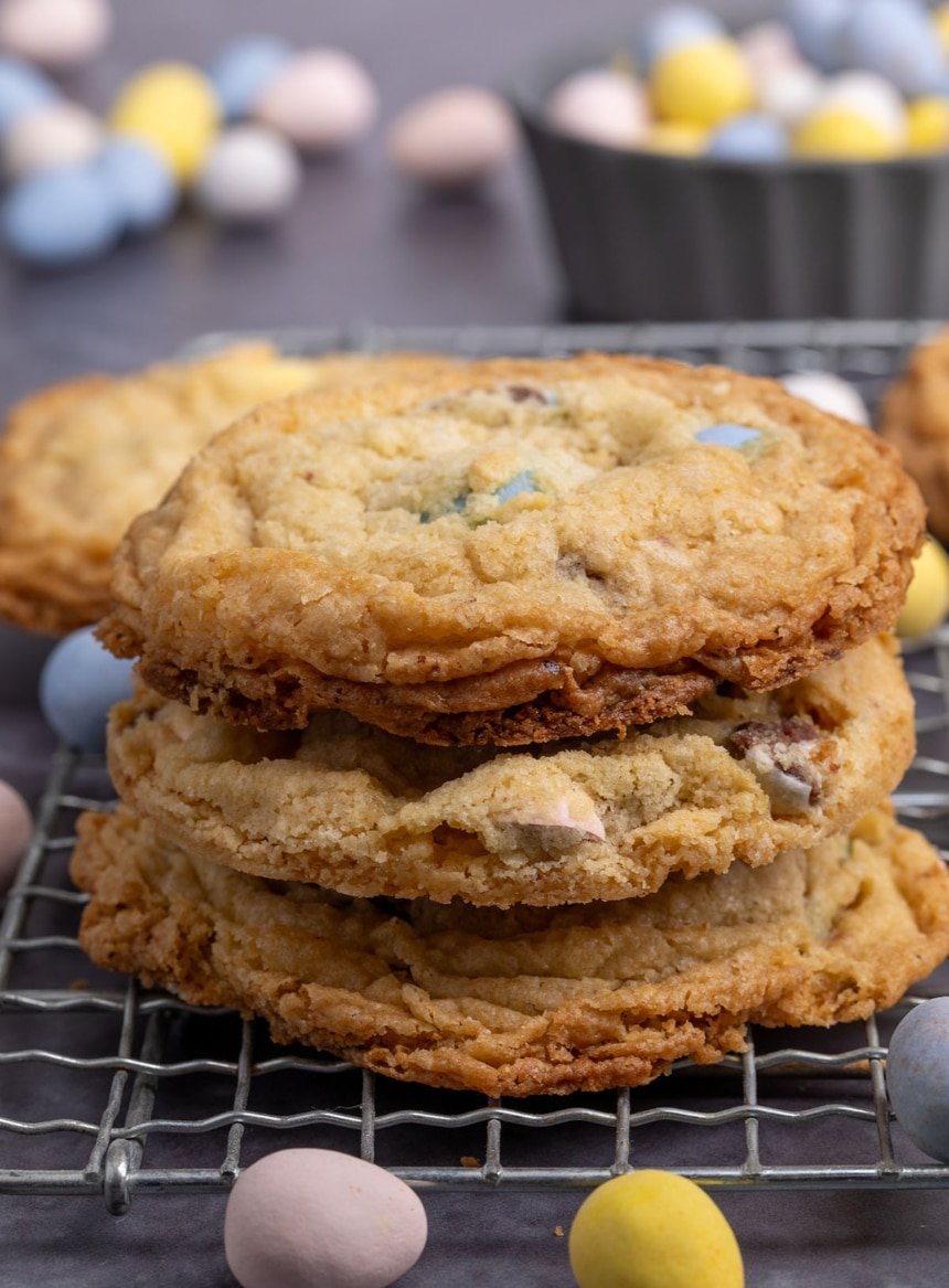 stack of 3 golden brown cookies pictured from the side on a metal wire rack with yellow and pink Cadbury mini egg candies on the side and some peeking out from the cookies