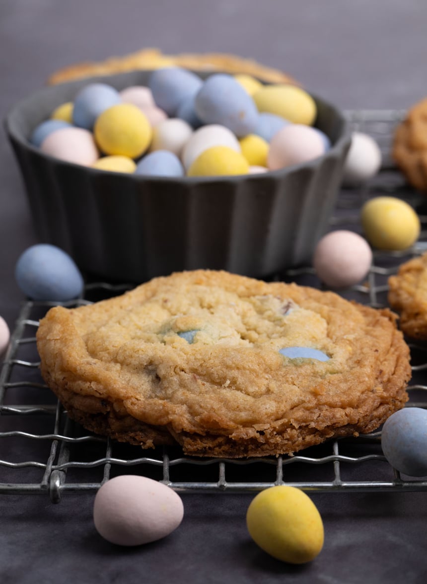 side view of one golden brown baked cookie with blue candy pieces peeking out and other candies on black surface nearby