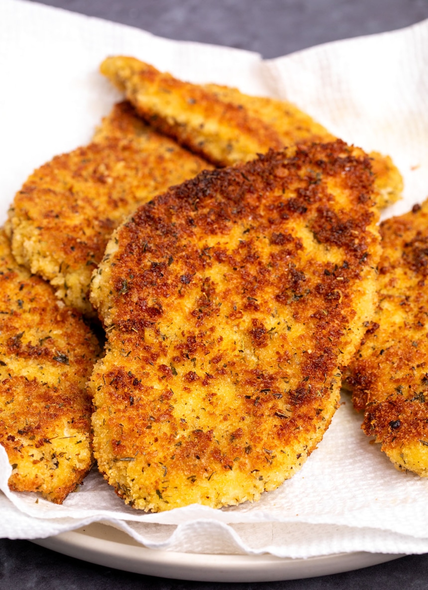 golden brown breaded and fried chicken cutlets with green herb flecks on white paper towels on light tan plate