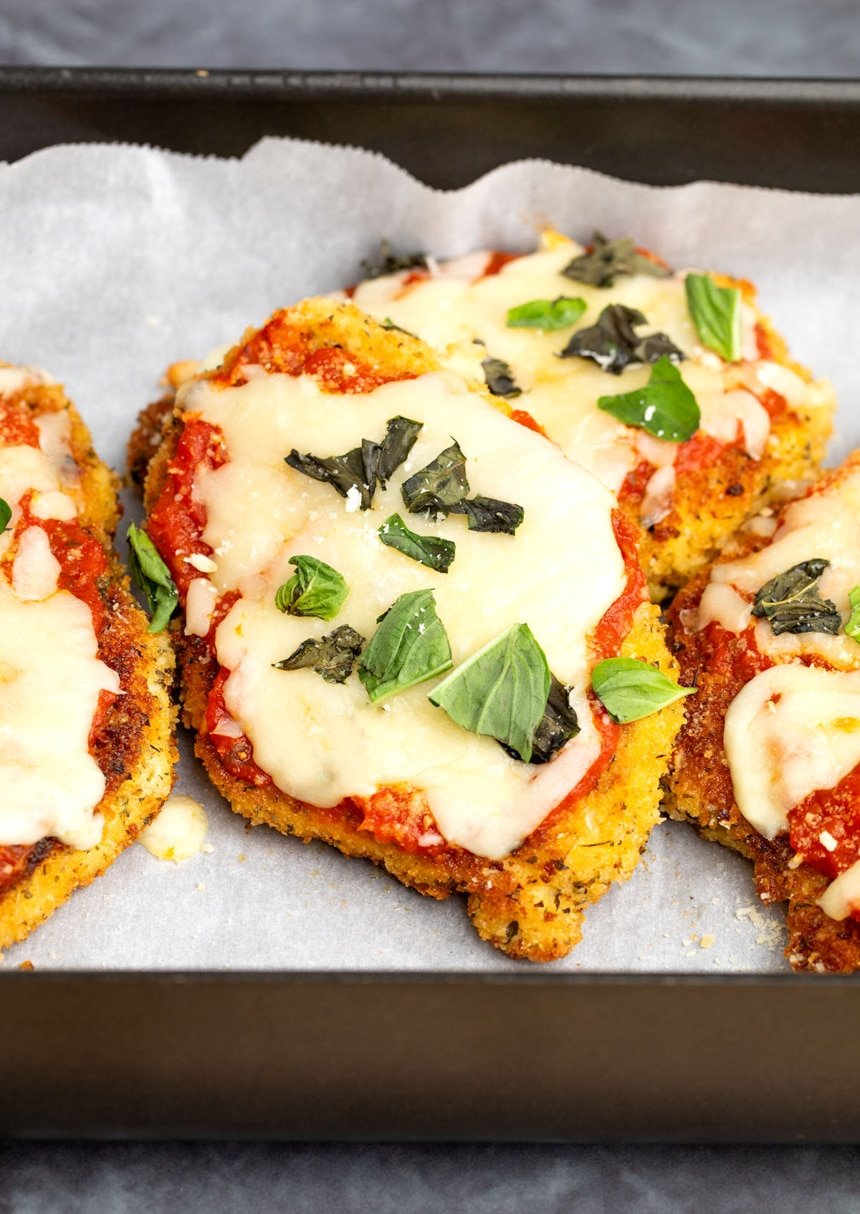 golden brown breaded chicken cutlets with red tomato sauce white melted cheese and green chopped basil on white paper in black casserole dish