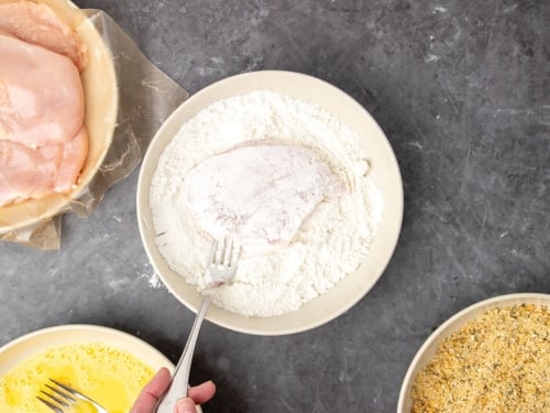 Fingers holding metal fork in wide flat light tan bowl with white flour and raw chicken cutlet covered in white flour