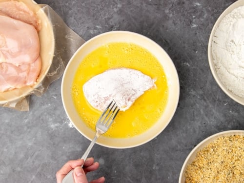 fingers holding metal fork dipping white flour-covered raw chicken cutlet into yellow beaten egg mixture in light tan bowl