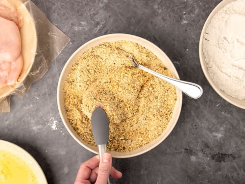 fingers holding gray silincone-tipped metal tons placing raw chicken cutlet in light brown breadcrumb mixture in flat bowl