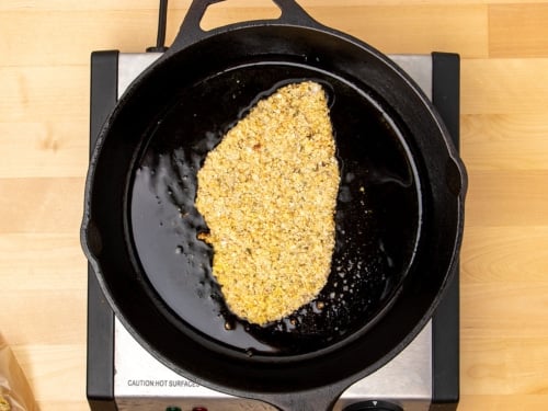 one raw breaded chicken cutlet in black round cast iron skillet cooking in bubbling oil