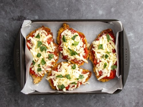 4 breaded chicken cutlets with tomato sauce, shredded mozzarella cheese and fresh basil in black casserole dish lined with white parchment paper