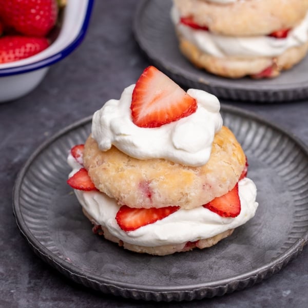 small round dark gray plate with light brown biscuit cut in half with white whipped cream and sliced red strawberries in the center and on top