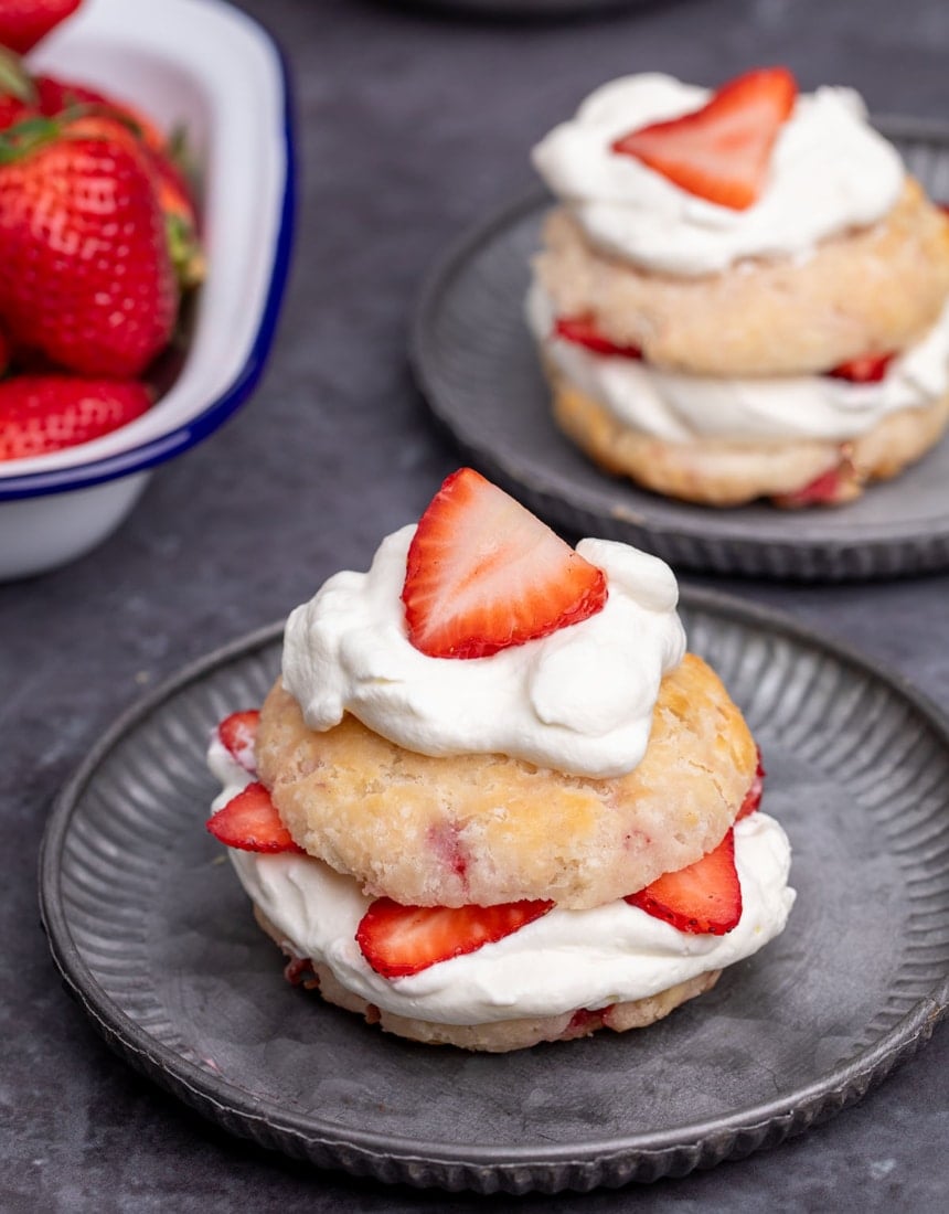 tan round biscuit with bits of red strawberries sliced in half horizontally with soft white whipped cream and sliced strawberries in the center and on top on a small dark gray plate with another similar plate and a white dish of red strawberries in the background