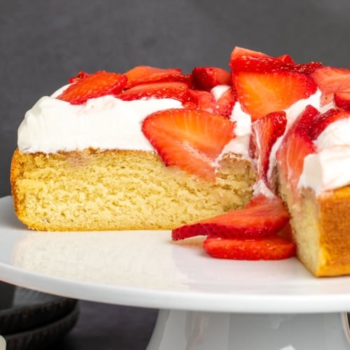 single layer yellow cake with 2 slices missing with white whipped cream and red sliced strawberries on white cake plate