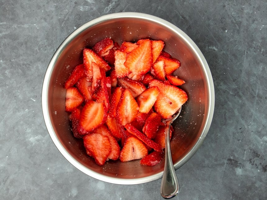 small round metal mixing bowl with sliced strawberries mixed with sugar with metal mixing spoon