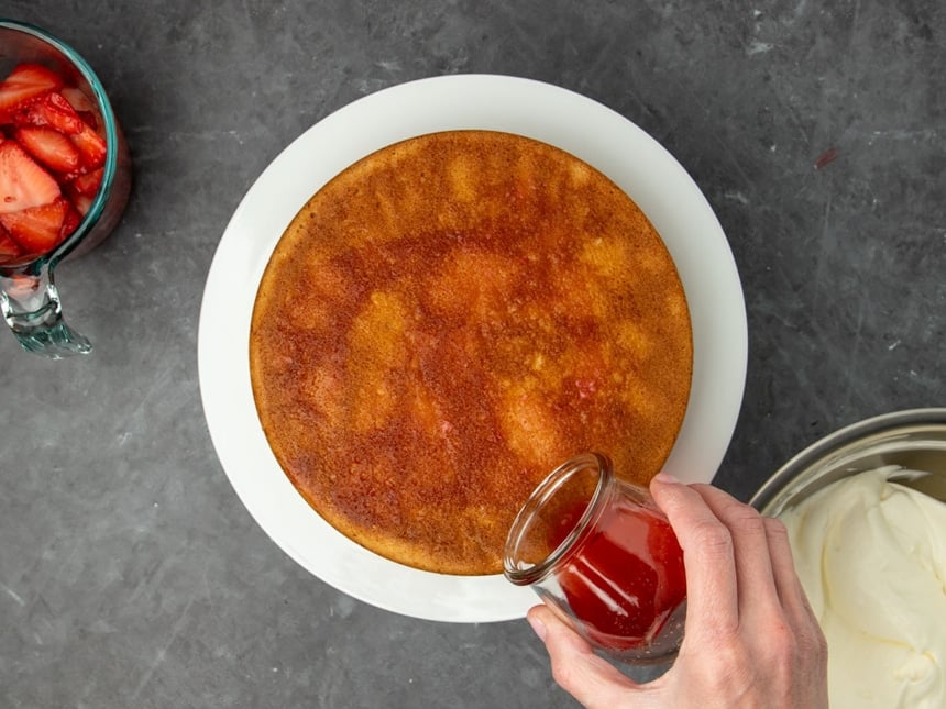 hand holding small glass jar pouring rendered strawberry liquid onto light brown round cake on white cake plate