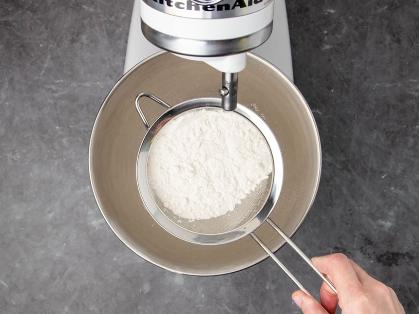 hand holding medium-size metal sieve with white flour over mixing bowl