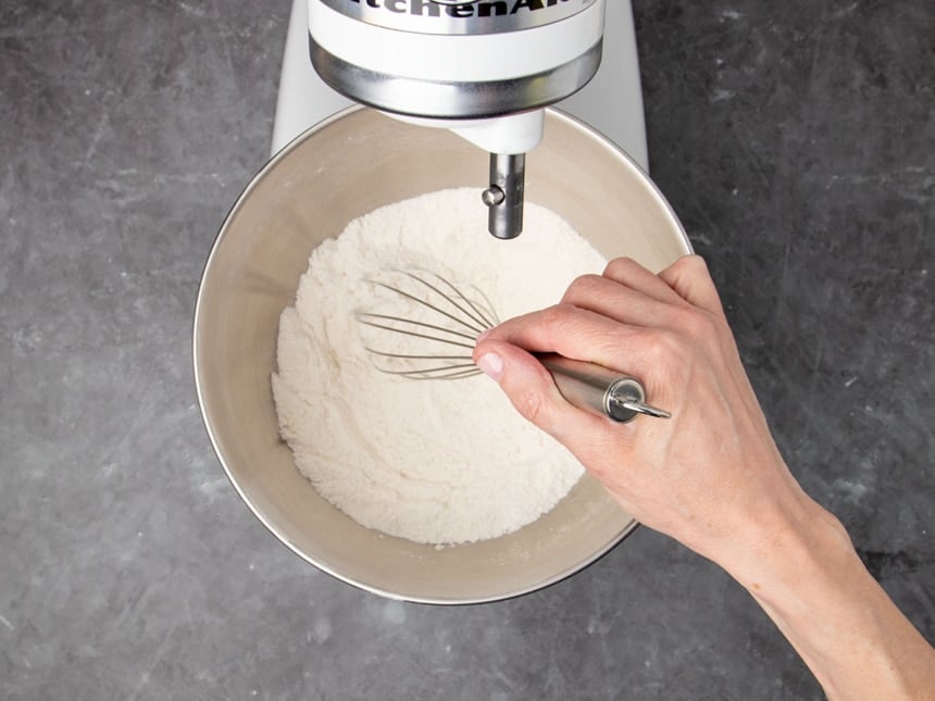 hand holding metal wire whisk, whisking white flour mixture in bowl of stand mixer