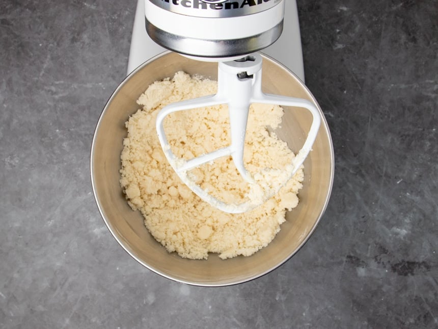 round metal bowl of stand mixer with clumping pale yellow flour and butter mixture with white mixer paddle