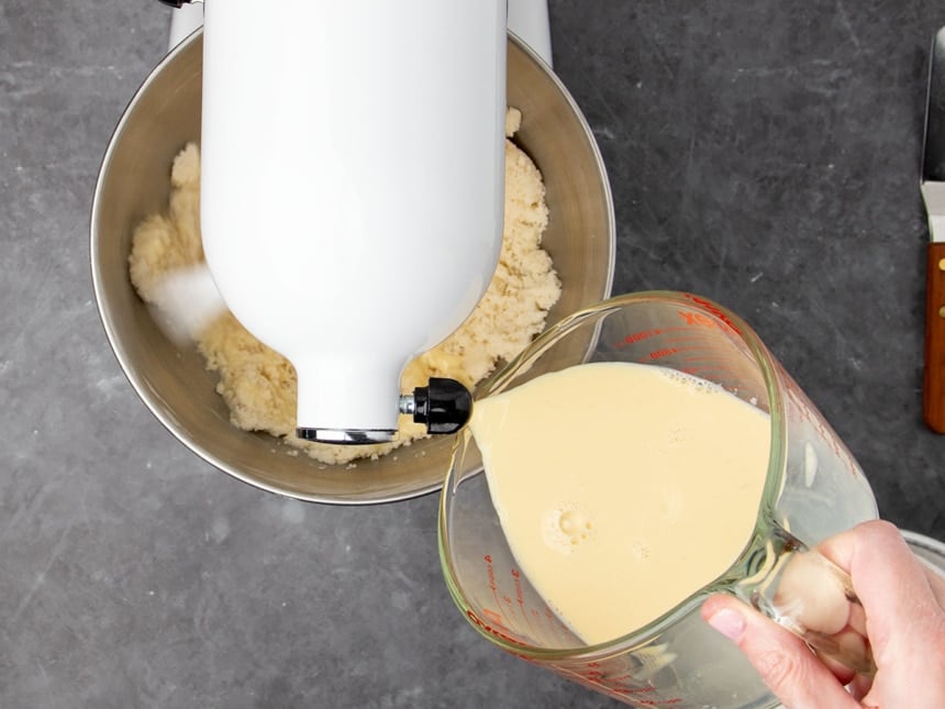 mixer on low speed while hand holding measuring cup pours yellow liquid into mixing bowl