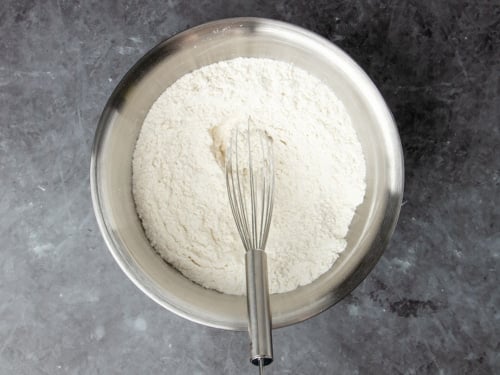 round metal mixing bowl with white flour mixture and large metal wire whisk