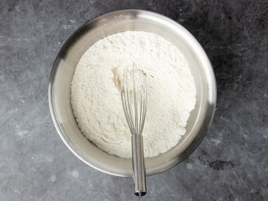 round metal mixing bowl with white flour mixture and large metal wire whisk