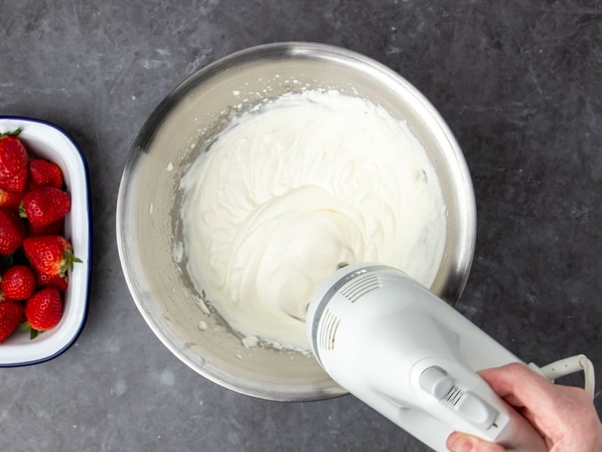 hand holding white handheld mixer being used to whip white cream into light and fluffy whipped cream in round metal mixing bowl with red strawberries in white dish on the side