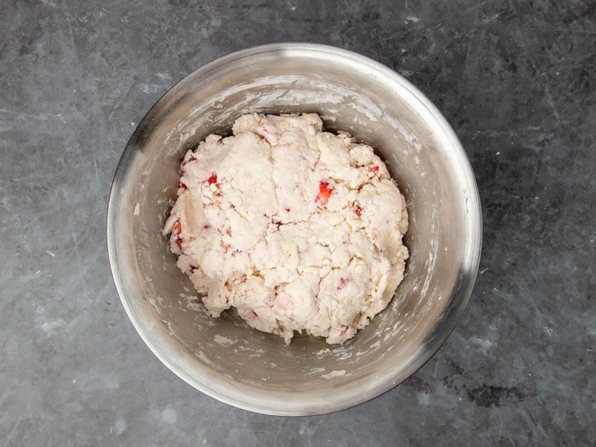 raw cream colored disk of raw biscuit dough with bright red bits of strawberries, still in mixing bowl