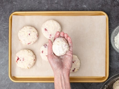 hand holding raw round mound of pale cream colored biscuit dough held above raw biscuits on white paper on gold colored baking pan