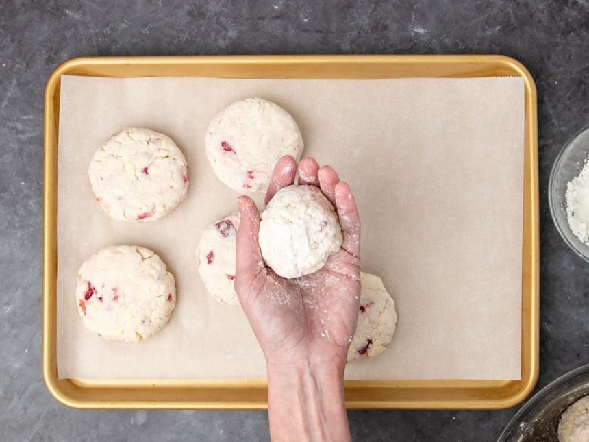 hand holding raw round mound of pale cream colored biscuit dough held above raw biscuits on white paper on gold colored baking pan