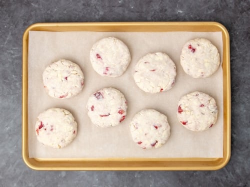 8 raw disks of cream colored biscuit dough with bright red specks of chopped strawberries on white paper on gold baking tray