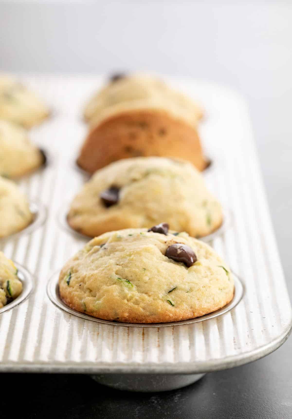 Light brown zucchini muffins with green flecks and brown chocolate chips in a metal muffin tin.
