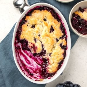 oval white baking dish with brown topping and blue blueberry filling with some of the cobbler missing from the dish