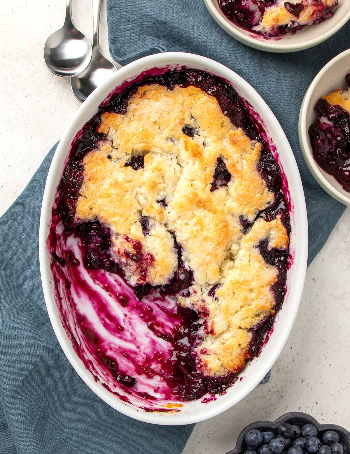 two metal spoons next to oval white serving dish with brown topping over purple and blue blueberry filling with more blueberries and servings in 2 small sand colored bowls on blue cloth