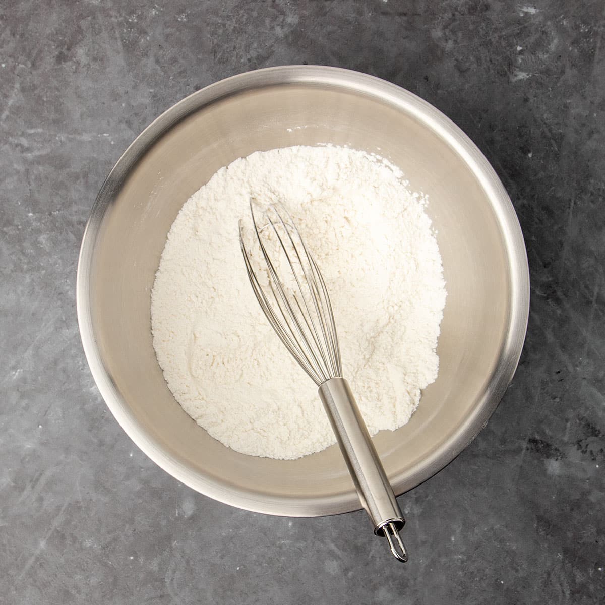 Large round metal mixing bowl with white flour mixture and metal wire whisk